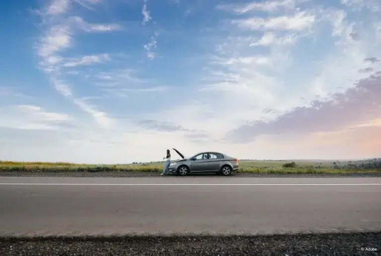 From across the road, a grey car with an open hood parked on the side of the road. A woman stands in front of the vehicle.