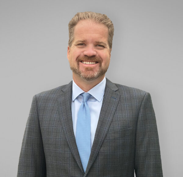 A professional headshot of Brian Plummer, with a grey suit and a blue tie.