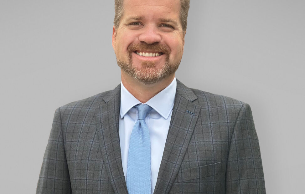 A professional headshot of Brian Plummer, with a grey suit and a blue tie.